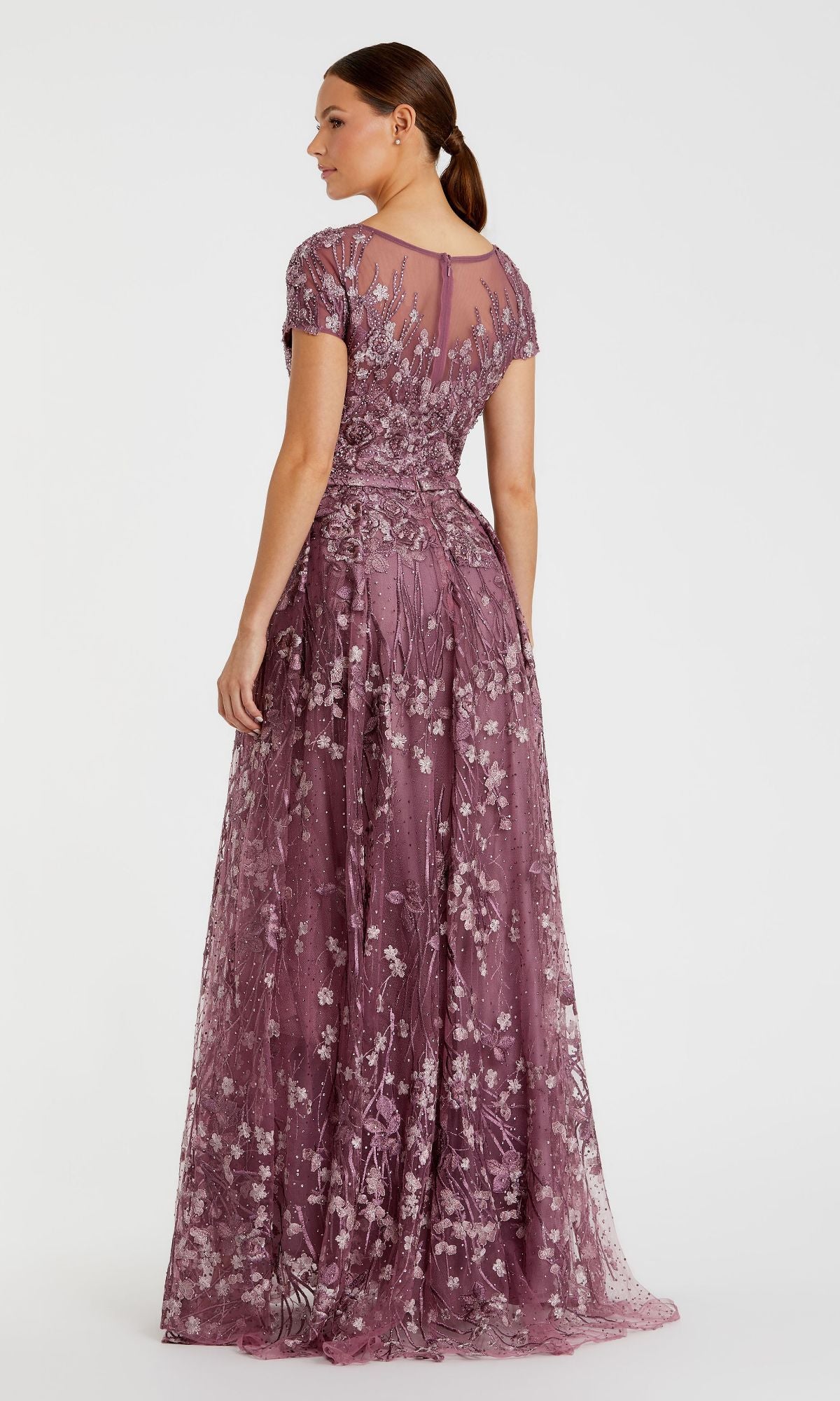 A woman wears the Mac Duggal 20405, a mauve short-sleeve long formal dress with embroidered lace, floral details, and a sheer back panel, standing against a plain white background.